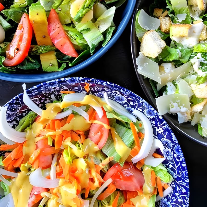 Three vibrant salads, including a Caesar salad, with fresh vegetables and dressings in various bowls.