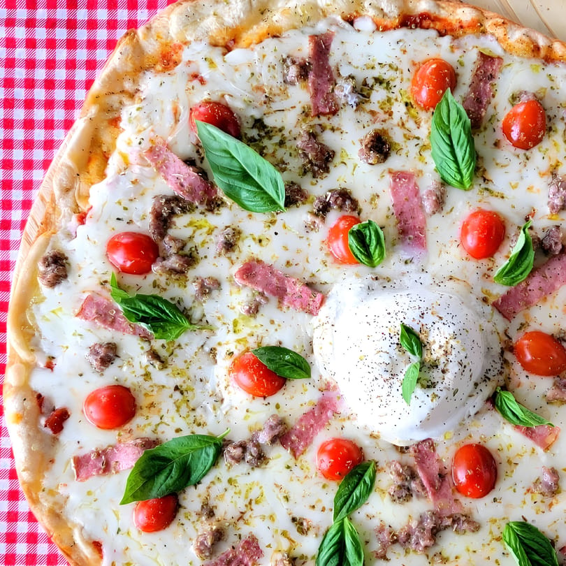 La Piccolina pizza with burrata, cherry tomatoes, basil, sausage, and prosciutto on a red checkered tablecloth.