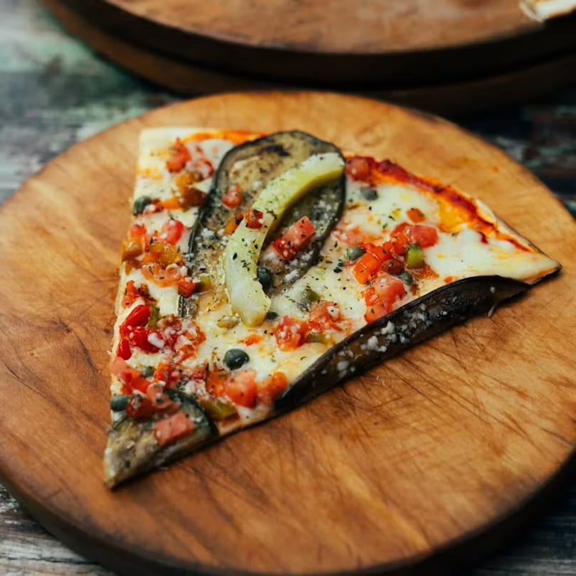 A slice of La Piccolina's pizza with eggplant, tomatoes, and cheese on a wooden board.